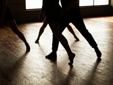 Nico S. Rubio coaching dancers on a wooden floor, warm window light filtering through, focus on feet in motion, 35mm film style.