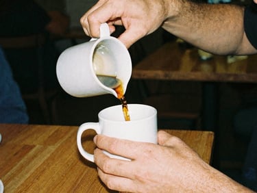 Close-up of hands pouring coffee into a mug at a solidarity café, warm indoor ambient light, candid moment, analog film grain.