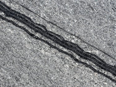 Close-up of Markino granite slab under raking natural daylight — mid-grey ground with bold black and white mineral banding, satin-polished surface, sharp shadow lines across the grain revealing textural variation
