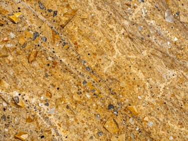 Close-up of Crystal Yellow granite slab under natural daylight — warm amber-gold ground with scattered dark mineral inclusions and white quartz veins, polished surface showing depth and warmth, raking light from the left