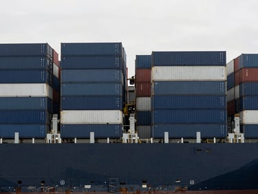 Wide shot of a cargo ship being loaded at a modern port, containers stacked geometrically, neutral overcast daylight, desaturated navy and gray tones, sharp architectural focus, 35mm lens, no people.