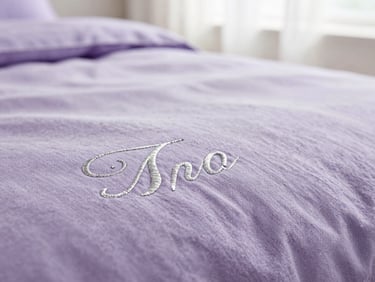 Close-up of soft lavender cotton fabric with delicate silver embroidered lettering, shallow depth of field