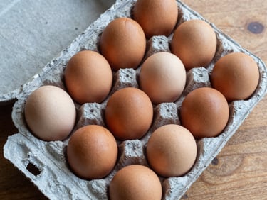 Overhead close-up of desi eggs in an open cardboard box, brown speckled shells with visible rough texture, placed on a wooden sorting table under natural overhead daylight, clearly distinct from standard eggs