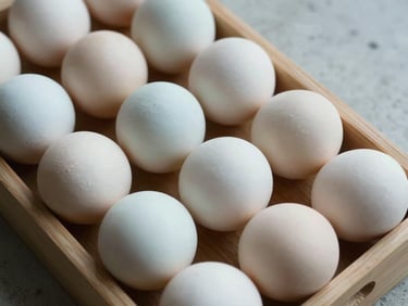 Overhead close-up of duck eggs lined in a shallow wooden tray, large off-white shells with a slight blue-grey tint, resting on a plain concrete surface under flat work-site lighting