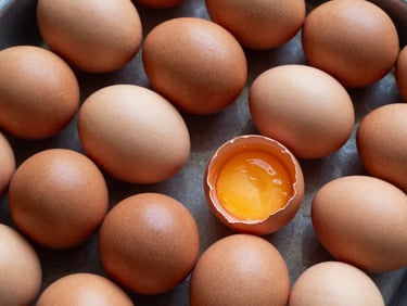 Close-up overhead shot of desi eggs in a sorting tray, brown and speckled shells with visible texture variation, natural daylight from above, a few eggs showing the darker yolk colour through a cracked shell on the side