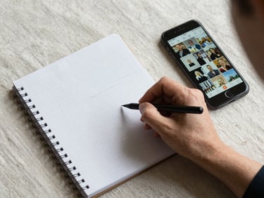 Overhead view of a planning session — a hand writing in a grid notebook on a pale linen surface, a phone showing an Instagram grid beside it, north-facing daylight, no artificial light