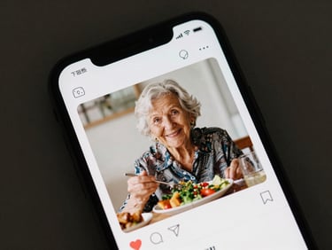Close overhead view of a smartphone screen showing a clean app feed with a photo of an elderly woman smiling at lunch, soft natural light, documentary framing, no staged pose