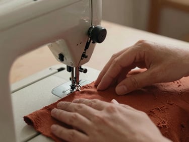Close-up of artisan hands guiding a piece of textured rust-colored cotton fabric through a vintage sewing machine, warm side lighting.
