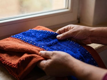 Close-up documentary photograph of hands sharing traditional embroidered textiles, warm window light, rich terracotta and lapis tones.