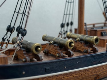 Macro close-up shot under warm desk light of a highly detailed wooden ship model rigging and brass cannons on a dark oak surface.