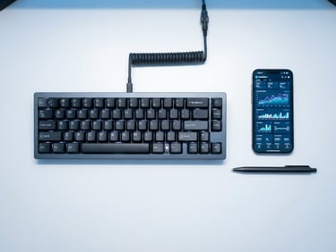 Top-down flat lay of a modern mechanical keyboard, a smartphone showing active analytics charts, and a sleek metallic pen on a clean white desk, illuminated by cool blue and white studio light.