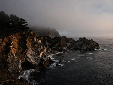 Moody coastal cliffs of Big Sur shrouded in thick fog at first light, high-contrast dark cliffs, deep charcoal water, cinematic lighting.