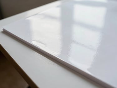 Close-up of coated gloss paper sheet held at a slight angle, smooth reflective surface catching diffuse window light, brilliant white finish, paper edge visible showing thin coating layer