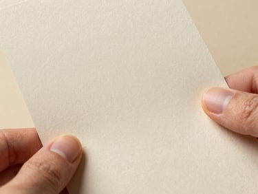 Close-up of specialty paper swatch held between two fingers, showing fine grain and subtle embossed texture, diffuse daylight, warm cream tones