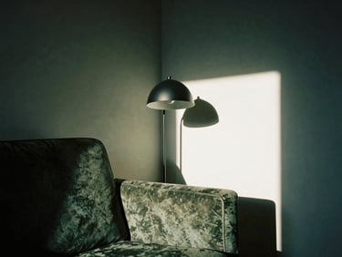 A sunlit corner of a living room, a worn velvet armchair next to a modern matte black steel reading lamp, casting deep shadows on a parchment wall, 35mm