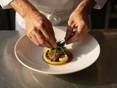 Tight overhead shot of a chef's hands plating a dish on a white ceramic bowl, golden-hour light raking across the counter, herbs and sauce visible mid-motion