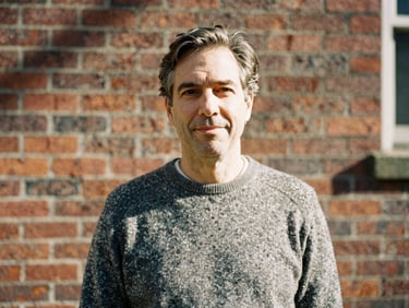 A warm editorial portrait of a middle-aged man in a wool sweater, soft daylight, outdoor historic brick wall background.
