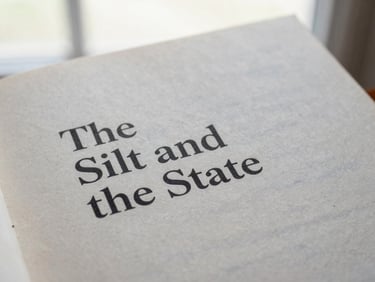 Close-up of a printed book cover titled 'The Silt and the State' resting on textured archival paper under soft, diffused window light, shallow depth of field, 35mm