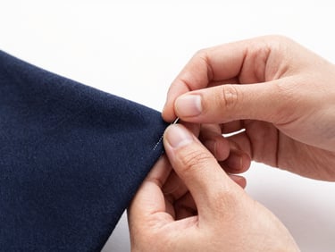 Close-up of a hand performing a blind hem stitch on dark navy wool fabric, crisp natural studio light, 35mm