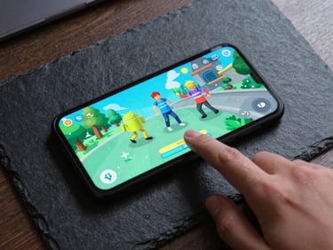 Overhead shot of a physical Android device on a dark slate desk, a finger tapping a colorful casual game interface with electric-cyan screen glow.