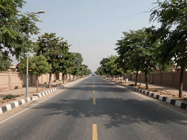 Ground-level architectural photograph of a wide asphalted road inside WAPDA Town Taru Jabba, plot survey markers and boundary walls visible on both sides, mature trees beginning to line the road, clear daytime light, framed from road center looking straight ahead toward the planned grid interior