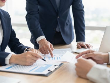 Modern office environment, collaborative meeting, hands analyzing reports, corporate navy and warm neutral tones, bright natural daylight, shallow depth of field, 35mm