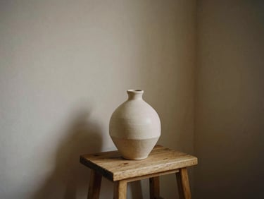 A minimalist interior corner with a raw ceramic vase on a wooden stool, soft shadows, warm-toned editorial photography, 35mm film