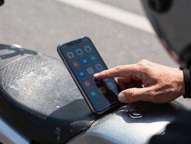 Tight landscape shot of a rider's thumb tapping a phone screen showing multiple service icons, shot from above on a worn motorbike seat, strong midday window light, pavement visible in shallow background focus