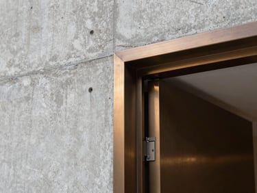Architectural interior detail showing the junction of a raw concrete wall and a brushed bronze door frame, precise craftsmanship, soft natural light.