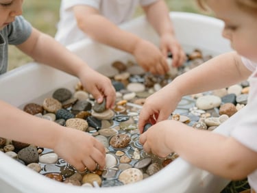 Toddlers playing with colorful sensory bins filled with water and natural stones, shallow depth of field.