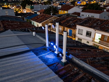 Overhead view of Colombian small-town rooftops with visible network infrastructure cables, sharp white and blue ambient light, precise technical composition