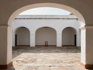 Wide environmental interior of a whitewashed courtyard seen through a low archway, late-morning light pooling on uneven flagstone, a single wooden chair in the middle distance
