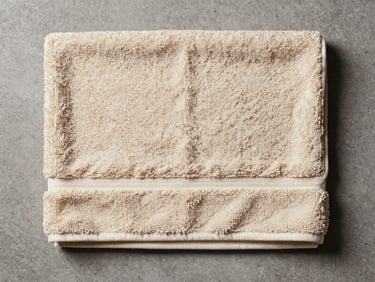 Flat-lay overhead shot of a folded bath towel on a grey concrete surface, diffused studio strobe lighting, close enough to show cotton terry loop density and hemline stitching, warm ivory tones, no props