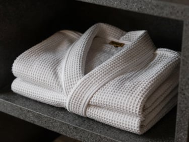 Close-up of a spa robe folded on a dark grey stone shelf, studio strobe lighting picking out the waffle-weave texture and a woven label on the collar, no people, tight framing