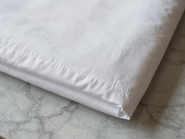 Overhead flat-lay close-up of a neatly folded hotel bed sheet corner on a marble surface, diffused daylight showing 400 thread count weave clarity and crisp hem stitching, no people