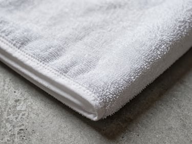 Extreme close-up of a hotel hand towel corner on a grey concrete surface, tight studio strobe lighting revealing the looped terry weave texture and a faint embroidered border detail, no people, overhead angle