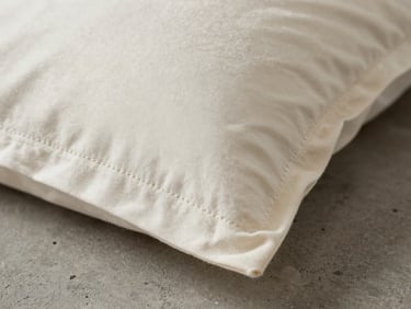 Extreme close-up of pillowcase corner with visible embroidery stitch line on ivory cotton, photographed under studio strobe on a grey concrete surface. Thread precision in sharp focus. No faces, no props.
