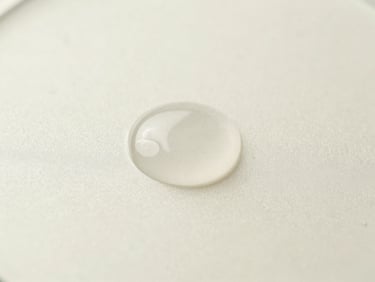 Macro close-up of a clear, viscous serum droplet on a frosted glass surface, soft clinical daylight, muted cream background