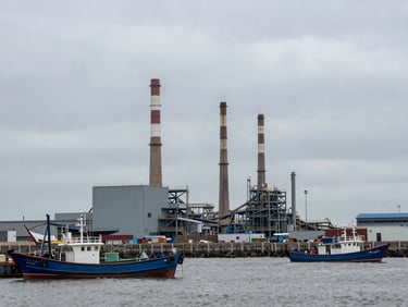 Wide environmental shot of a coastal industrial zone at overcast midday, smokestacks visible against flat grey sky, fishing boats moored in the foreground