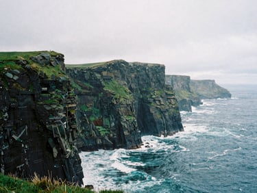 Dramatic panoramic shot of the Herbeira cliffs, misty and rugged, under an overcast sky, with crashing waves far below, moody coastal photography, deep green and granite tones, 35mm film