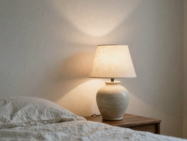 A ceramic bedside lamp casting warm soft light on a textured plaster wall, linen bedding nearby