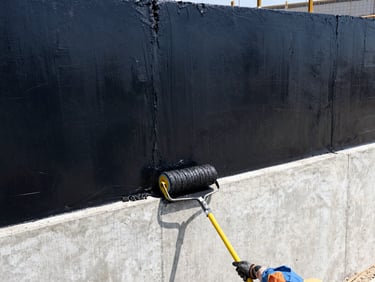 Worker applying liquid black bitumen coating to a concrete retaining wall with a roller, high contrast daylight, architectural construction details