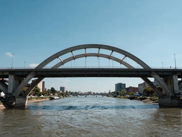 Head-on ground-level view of a wide concrete arch bridge over a major river, sharp midday light casting strong shadow under the arch, clear blue sky, urban context visible at both banks