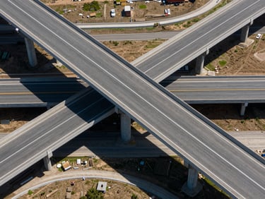 Aerial view of a completed highway interchange with multiple elevated ramp structures, photographed straight down from above, midday sun, precise geometric shadows of concrete piers on asphalt below