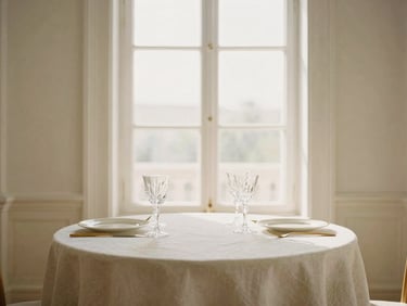 Warm editorial photography of a minimalist luxury table setting, fine linen tablecloth, gold cutlery, delicate crystal glassware, soft afternoon window light, 35mm