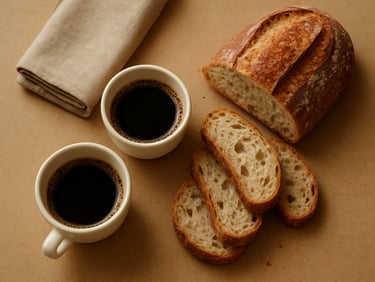 Close-up overhead of artisanal coffee-break spread — small cups, sliced pão artesanal and a folded linen napkin on a warm beige surface, soft diffused daylight, no people