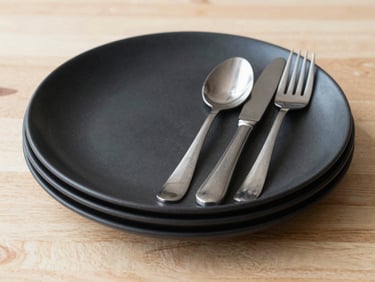 Matte black ceramic plates and stainless steel cutlery stacked on a light timber tabletop, straight-on architectural crop