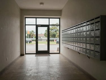 Wide shot of a building entrance hallway with tiled floor and mailboxes on the right wall, overcast daylight from glass door ahead, no people, straight-on framing