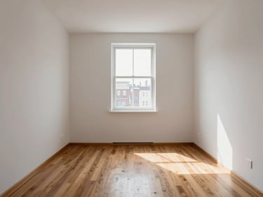 Straight-on interior shot of a compact city apartment living room under north-facing window light, white walls, hardwood floor, empty space, no décor