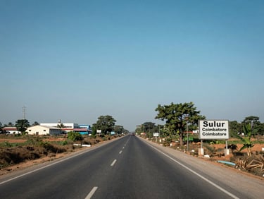 Wide environmental daylight photograph looking down a straight road in Sulur leading toward Coimbatore — a signboard for a school visible on the right roadside, industrial buildings in the mid-distance on the left, clear blue sky, low-angle framing that emphasises the road extending toward the city, no people